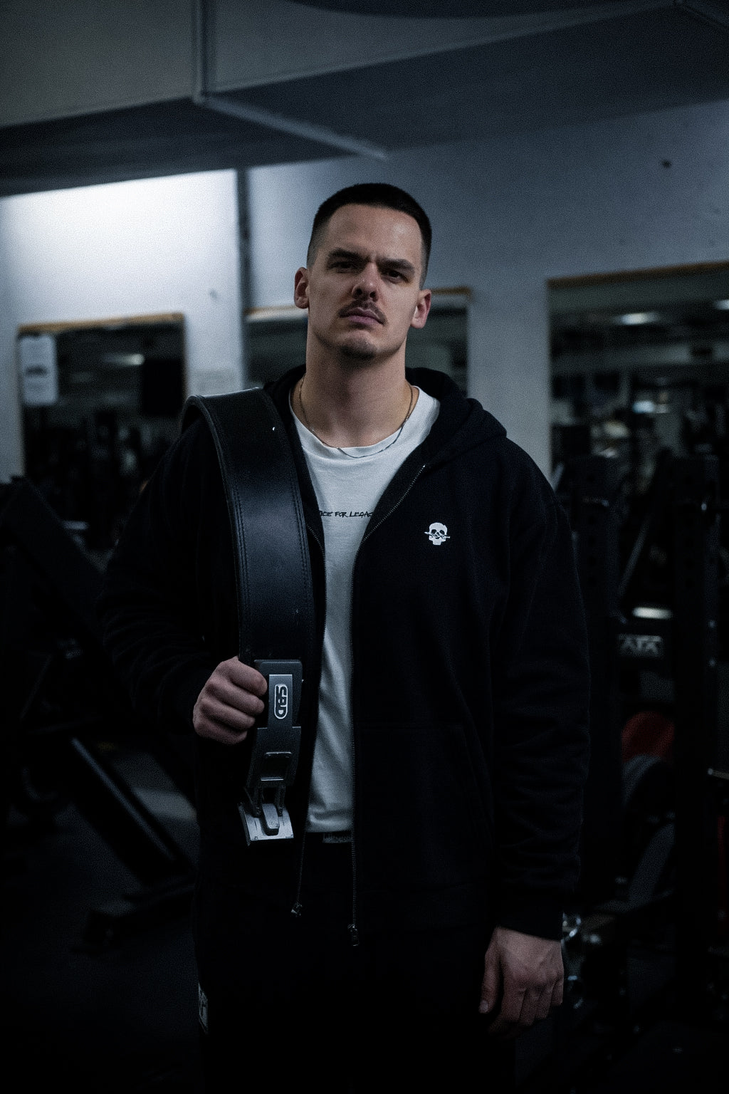 Athlet mit SBD-Gewichthebergürtel über der Schulter im schwarzen Azan Strength Zip-Hoodie im Gym